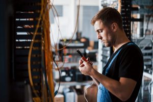 Young man in uniform with measuring device works with internet equipment and wires in server room.